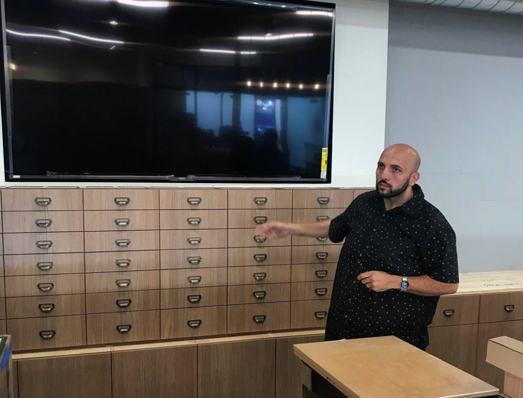 Interior view of man standing and gesturing in front of a bank of small drawers that have a large dark monitor above.